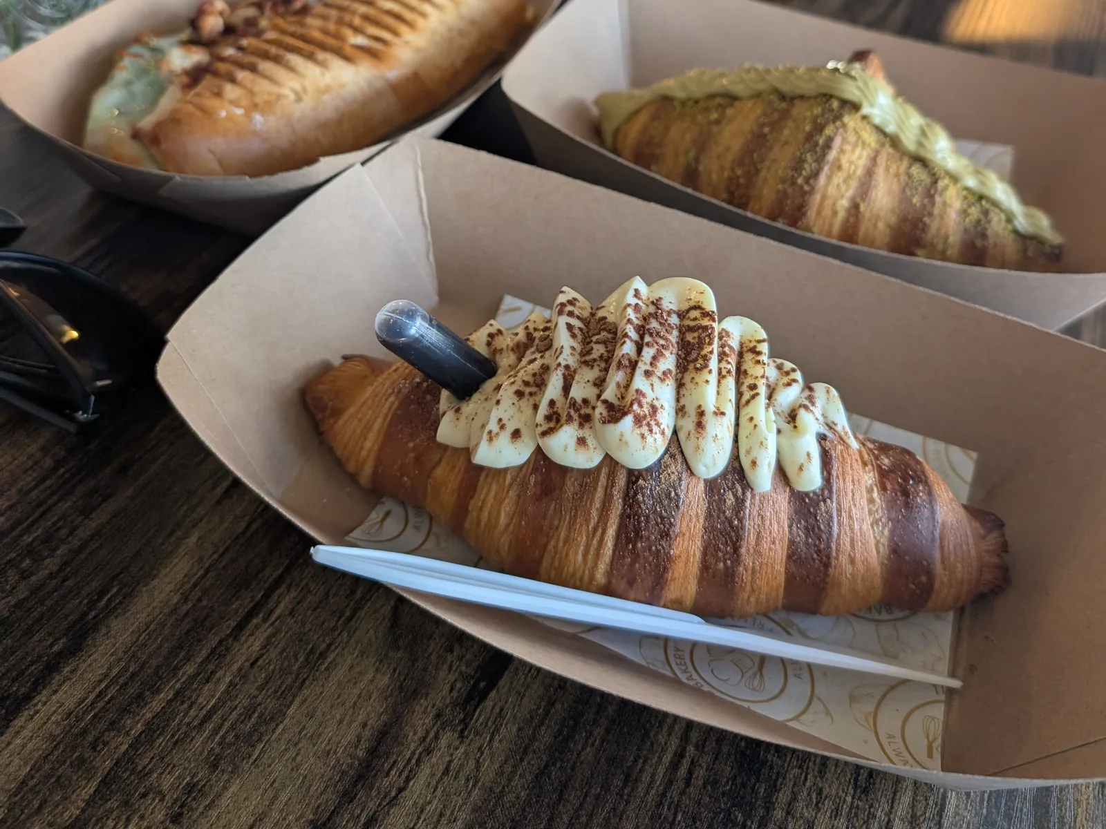 A pistachio croissant piped with cream and dusted with crushed pistachio, in a paper tray beside two toasted sandwiches at Tico's Bakery in Tampa