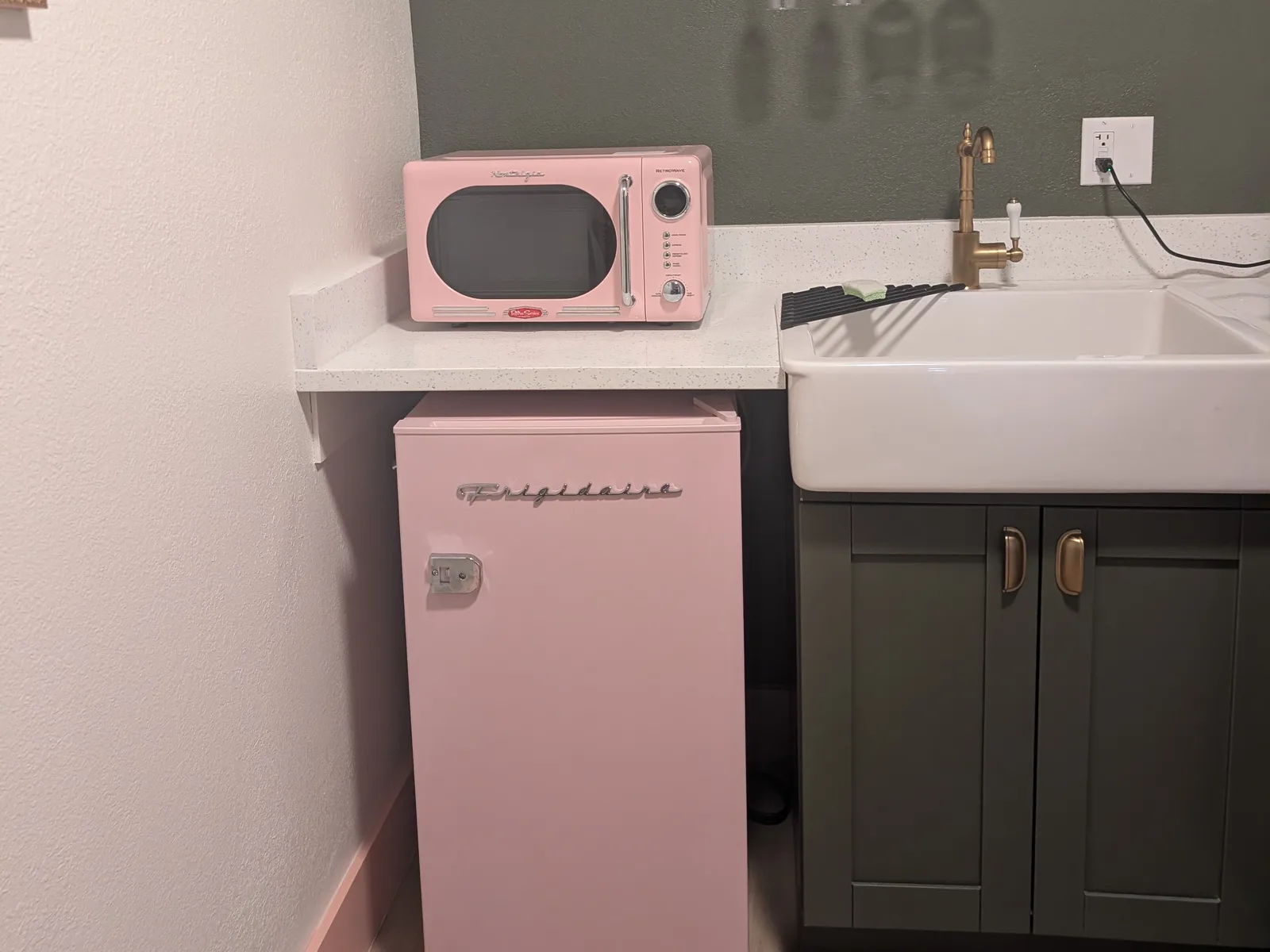 A retro kitchenette in a suite at Abode Beach House: a pink microwave and a pink Frigidaire fridge beside a farmhouse sink with a brass tap and dark green cabinets